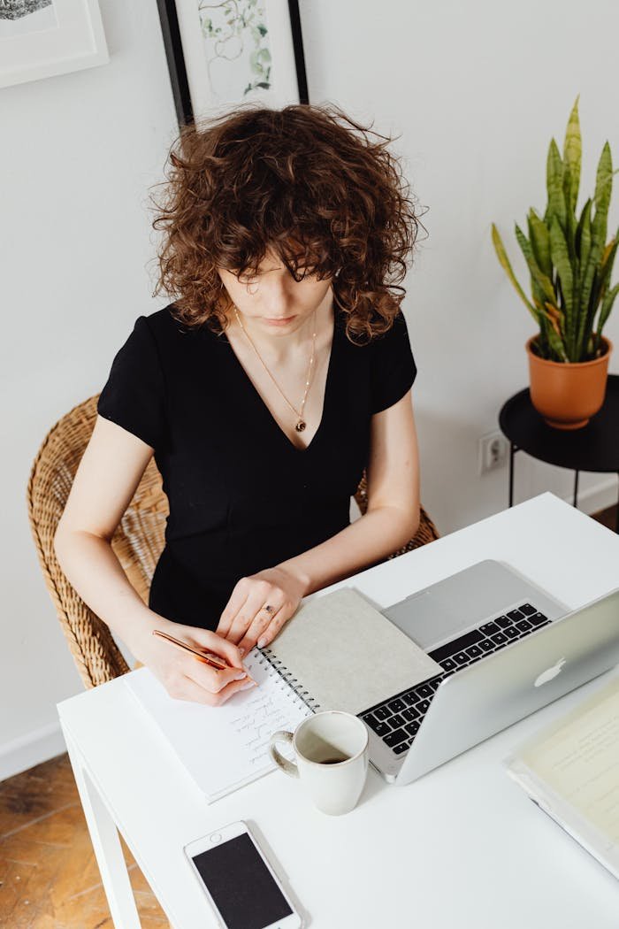 High-Angle Shot of a Woman Writing on a Notebook Near a Laptop